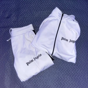 Vit track jacket från Palm Angels - Vit track jacket från Palm Angels med svarta detaljer och dragkedja framtill. Jackan har svarta ränder längs axlarna och ärmarna, hög krage och logga på bröstet. Tillverkad i ett glansigt syntetmaterial som ger en sportig vibe.