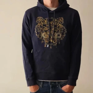 Mörkblå Kenzo hoodie med tigertryck - Snygg mörkblå hoodie från Kenzo med ikoniskt tigertryck i guld på bröstet och Paris-text. Tröjan har huva med snörning och en klassisk känguruficka framtill. Perfekt för dig som vill sticka ut med ett coolt motiv.