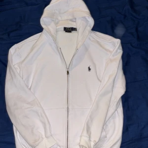 Vit hoodie från Polo Ralph Lauren - Vit hoodie från Polo Ralph Lauren i bomull med klassisk broderad logga på bröstet. Tröjan har huva, dragkedja framtill och långa ärmar med ribbade muddar. Snygg och enkel design som passar till många olika stilar.