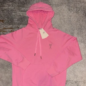 Rosa hoodie från Ami med hjärtdetalj - Säljer en snygg rosa hoodie från Ami med huva och snörning. Tröjan har ett broderat hjärta med A på bröstet och klassisk känguruficka framtill. Perfekt för dig som gillar streetwear och vill sticka ut med färg. Taggarna sitter kvar.