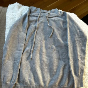 Grå hoodie med snörning - Snygg grå hoodie med långa ärmar och snörning i huvan. Tröjan har ribbade muddar vid ärmslut och nederkant, perfekt för en chill och avslappnad stil. Materialet känns mjukt och skönt mot huden.