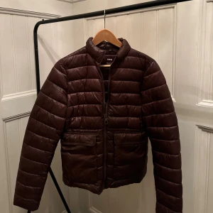 Brun pufferjacka från MNG, storlek L - Snygg brun pufferjacka från MNG i storlek L. Jackan har quiltad design, två stora fickor med lock och knapp framtill samt dragkedja. Perfekt för kyliga dagar med sin lätt vadderade känsla och stilrena look.