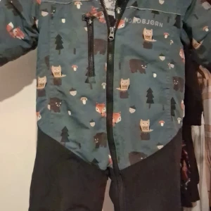 Nordbjørn vindjacka med djurmönster - Säljer en grön vindjacka från Nordbjørn med sött djurmönster som inkluderar ugglor, rävar och björnar. Jackan har svart dragkedja framtill och detaljer i svart på nedre delen. Perfekt för dig som gillar natur och roliga prints.