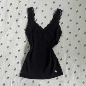 Svart spetslinne från Abercrombie & Fitch - Snyggt svart linne från Abercrombie & Fitch med breda axelband och spetsdetaljer längs v-ringningen. Ribbad struktur och liten vit logga nertill. Perfekt för dig som gillar stilrena och feminina toppar.