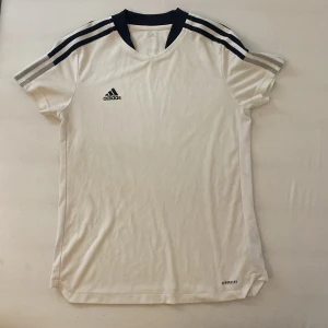 Vit Adidas fotbollströja M - Snygg vit fotbollströja från Adidas i storlek M, tillverkad i Primegreen-material. Tröjan har svarta detaljer vid kragen och axlarna samt klassiska Adidas-ränder på ärmarna. Kortärmad och med AEROREADY-teknologi för extra komfort under träning.
