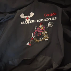 Svart Moose Knuckles pufferjacka L - Svart pufferjacka från Moose Knuckles med stor fluffig huva och snygga silverfärgade dragkedjor. Jackan har broderad logga och coolt motiv på insidan, samt flera fickor med dragkedja. Perfekt för kalla dagar och har en riktigt urban vibe.