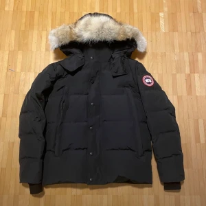 Svart dunjacka från Canada Goose - Svart dunjacka från Canada Goose med fluffig päls på huvan och klassisk logga på ärmen. Jackan har både dragkedja och knappar framtill, samt ribbade muddar vid ärmslut. Perfekt för kalla vinterdagar och har en stilren, boxig passform.