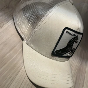 Vit keps The Farm Goorin Bros Stallion - Snygg vit keps från The Farm Goorin Bros med mesh på sidorna och bak, samt broderad patch med svart häst och texten 'Stallion' framtill. Justerbar passform och böjd skärm. Perfekt för dig som gillar streetstyle och unika accessoarer.