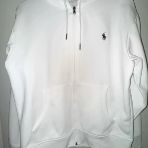 Vit hoodie från Polo Ralph Lauren - Vit hoodie från Polo Ralph Lauren med dragkedja, huva med snörning och två stora fickor framtill. Klassisk Polo-logga broderad på bröstet. Skön och mjuk i bomullsmaterial, perfekt för chill dagar. Passar dig som gillar enkel och stilren look.