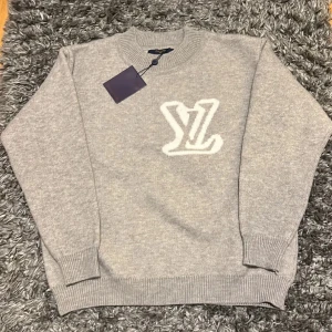 Grå Louis vuitton tröja - Exklusiv grå stickad tröja från Louis Vuitton i storlek S. Tröjan har ett stort vitt LV-logo framtill, rund hals och ribbade muddar vid ärmslut och nederkant. Perfekt för dig som vill ha en clean och lyxig look med designer vibe.