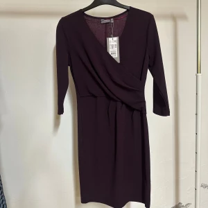 Mörklila omlottklänning från b.young - Supersnygg mörklila omlottklänning från b.young med v-ringning och trekvartsärm. Klänningen har ett draperat liv som ger en cool siluett och är tillverkad i mjukt polyester-material. Perfekt för dig som gillar stilrena och trendiga plagg med lite edge. Prislappen kvar