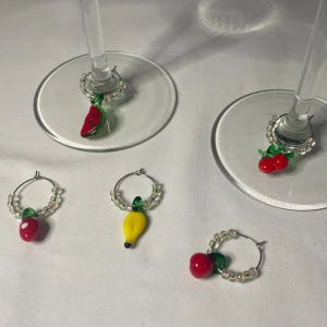 Glasmarkörer med fruktberlocker och pärlor - Fem markörer med färgglada fruktberlocker: äpple, körsbär, banan, vattenmelon och jordgubbe. Varje berlock har små pärlor på ringen och berlockerna är i glas med starka färger som röd, grön och gul. Perfekt för dig som gillar att förfina middagar och samtidigt hålla reda på glaset. 
