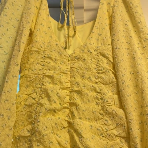 Gul blommig kortklänning från Nelly - Superfin gul kortklänning från Nelly med små blommor i grönt och vitt. Klänningen har långärmade puffärmar, snygg rynkning framtill och knytning i ryggen. Materialet är lätt och krinklat, perfekt för sommarens alla tillfällen. Söt och trendig look! Helt ny. Köpte i stl 36 och 38, men  glömde att lämna tillbaka. 