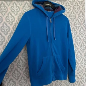 Blå hoodie från Burberry - Säljer en blå hoodie från Burberry med dragkedja och huva. Insidan av huvan har rutigt foder i blått, vinrött och svart. Tröjan har två fickor framtill och broderad Burberry-logga på ärmen.