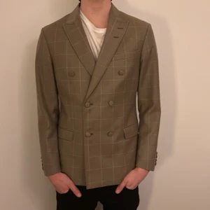 Beige rutig kavaj Tiger of Sweden - Snygg dubbelknäppt beige kavaj från Tiger of Sweden med diskret rutigt mönster. Klassisk passform med breda slag, fickor med lock och knappar framtill. Tillverkad i exklusiv ull från Marlane Super 100’s. Perfekt för dig som vill ha en stilren och modern look.HELT NY ALDRIG ANVÄND och PRIS KAN DISKUTERAS