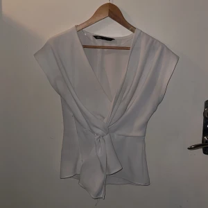 Vit omlottopp med knyt från Zara - Snygg vit omlottopp från Zara i storlek XS. Toppen har v-ringad hals, korta ärmar och ett knyt framtill som ger en cool detalj. Materialet är mjukt och lätt, perfekt för ljusa dagar. Dragkedja i sidan för enkel påklädning. Stilren och fräsch look.