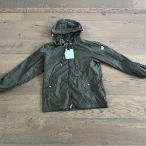 Grön regnjacka från Moncler - Snygg grön regnjacka från Moncler med huva och coola snörningsdetaljer. Jackan har dragkedja framtill, två fickor med tryckknappar och Moncler-logga på ärmen. Röd, vit och blå rand på huvan ger extra stilpoäng. Perfekt för dig som gillar streetwear.