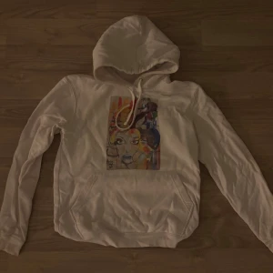 Vit hoodie med konsttryck - Vit hoodie med färgstarkt konsttryck framtill som föreställer ett ansikte. Tröjan har huva med snörning och en stor magficka. Tillverkad i mjuk bomull och har långa ärmar. Perfekt för dig som gillar unika detaljer och streetstyle.