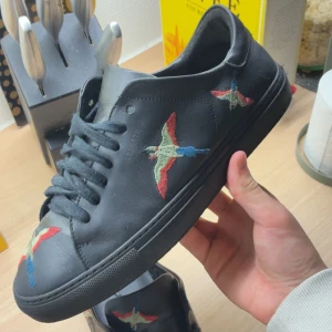 Svarta sneakers från Axel Arigato - Svarta sneakers i skinn från Axel Arigato med broderade fåglar i rött, blått och beige på sidorna och tån. Skorna har svarta snören och en klassisk låg profil. Perfekta för dig som vill ha en stilren men unik look.