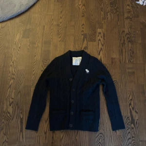 Svart kofta från Abercrombie & Fitch - Svart kabelstickad kofta från Abercrombie & Fitch med klassisk v-ringning och knappar framtill. Tröjan har två fickor och en vit logotyp med älg på bröstet. Perfekt för lager-på-lager och chill stil under höst och vinter.