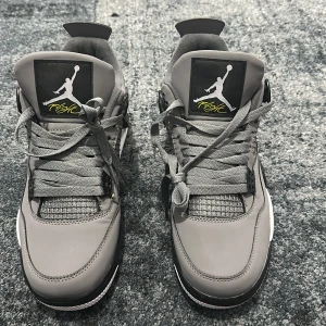 Nike Air Jordan 4 grå sneakers - Säljer ett par Nike Air Jordan 4 sneakers i grått med svarta och vita detaljer samt gula inslag på sula och tunga. Skorna har snörning, platt sula och rund tå. Ikonisk Jumpman-logga syns både på tungan och hälen. Ovandelen är i syntet och skinn för en clean look.