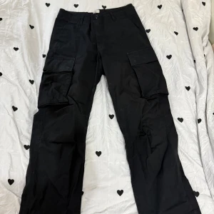 Svarta cargopants från H&M, strl 30 - Svarta cargopants från H&M med relaxed fit och stora fickor på sidorna. Byxorna har en rak passform och är tillverkade i bomull. Perfekta för dig som gillar streetwear och vill ha mycket plats för prylar.