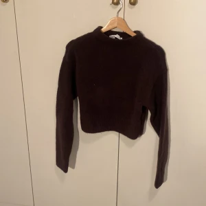 Mörkbrun croppad stickad tröja - Mysig mörkbrun stickad tröja från Zara med croppad passform och långa ärmar. Tröjan har en enkel design med ribbade muddar vid ärmslut och nederkant. Perfekt för kyliga dagar och enkel att styla med jeans eller kjol.