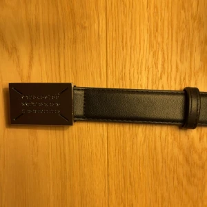 Svart bälte Maison Margiela - Säljer ett svart bälte från Maison Margiela med ikoniskt metallspänne där siffror är ingraverade. Bälten är i slätt skinn och har en stilren, minimalistisk design som passar till många outfits. Perfekt accessoar för dig som gillar exklusiva detaljer.