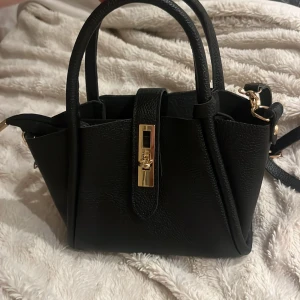 Svart handväska med gulddetaljer - Snygg svart handväska med guldfärgad låsdetalj framtill och två handtag. Väskan har en stilren design och är tillverkad i skinnimitation med struktur. Perfekt storlek för det viktigaste och har även en axelrem för extra flexibilitet.