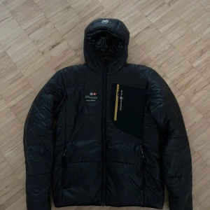 Svart pufferjacka från Sail Racing - Fon och fräsch Svart pufferjacka från Sail Racing med huva och gul dragkedja på bröstfickan. Jackan har en sportig look med logga på bröstet och quiltad design. Perfekt för kalla dagar och har en snygg, modern passform.