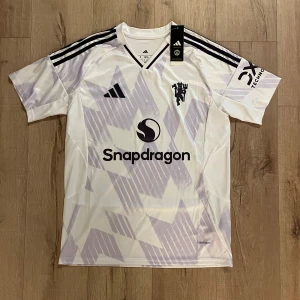 Manchester United vit bortatröja Adidas S - Manchester Uniteds bortatröja från Adidas i vit med ljuslila geometriskt mönster. Tröjan har svarta detaljer, V-ringad krage och korta ärmar med klassiska Adidas-ränder på axlarna. Materialet är lätt polyester med Aeroready-teknologi. Sponsorer: Snapdragon och DXC Technology.