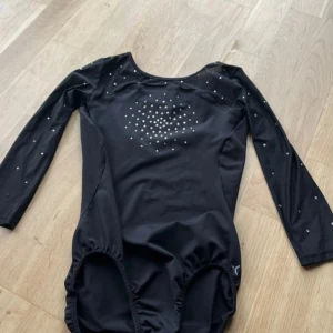 Svart långärmad gymnastikdräkt med stenar - Snygg svart gymnastikdräkt med lång ärm och glittriga stenar på bröstet och ärmarna. Dräkten har rundad hals och är tillverkad i stretchigt syntetmaterial som sitter bekvämt och följer kroppens rörelser. Perfekt för träning eller tävling inom gymnastik. Ärmarna ska vara lite kortare