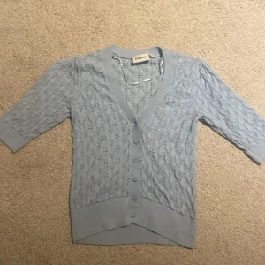 Ljusblå kofta från Kappahl - Söt ljusblå kofta från Kappahl med v-ringning och fina knappar framtill. Koftan har ett stickat spetsmönster och trekvartsärmar, perfekt för lager på lager. Ribbstickade muddar vid ärmslut och nederkant ger en snygg passform.