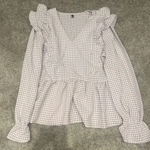 Rutig blus med volanger från Morango - Supersöt rutig blus från Morango i ljusgrått och vitt. Blusen har v-ringning, långa ärmar med volang vid ärmslut och volangdetaljer längs axlarna. Tillverkad i mjuk bomull, perfekt för dig som gillar romantiska och trendiga detaljer.