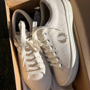 Vita Fred Perry sneakers med broderi storlek 38 herr aldrig använda - Snygga vita sneakers från Fred Perry med klassisk broderad lagerkrans i guld på sidan. Skorna har rund tå, platt sula och snörning med metallöglor. Tillverkade i bomullstyg med stilrena sömmar och diskret design. Perfekta för dig som gillar cleana och sportiga sneakers.