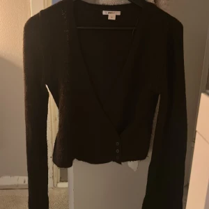 Mörk brun stickad kofta med v-ringning - Mörk brun stickad kofta från Gina Tricot med djup v-ringning och tre knappar framtill. Modellen är croppad och har långa ärmar, perfekt att slänga över en topp för en chill look.