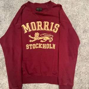 Vinröd sweatshirt från Morris XS - Snygg vinröd sweatshirt från Morris med stort gult tryck framtill där det står 'Morris Stockholm' och ett lejonmotiv. Tröjan har rund hals, långa ärmar och ribbade muddar. Perfekt för en chill och stilren look.