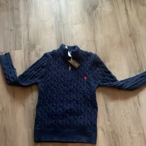 Mörkblå stickad tröja Polo Ralph Lauren - Mörkblå stickad tröja från Polo Ralph Lauren med klassiskt kabelstickat mönster och röd logga på bröstet. Tröjan har hög krage med half zip-dragkedja och långa ärmar. Perfekt för dig som gillar preppy och stilren look.