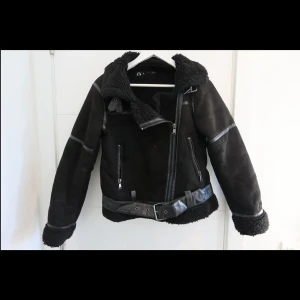 Svart teddyjacka från Zara, S - Svart teddyjacka från Zara i storlek S. Jackan har mjukt teddyfoder, detaljer i skinnimitation, två dragkedjefickor och bälte vid midjan. Snygga sömmar och grov krage ger en cool vibe. Perfekt för kalla dagar när du vill vara både varm och stilren.