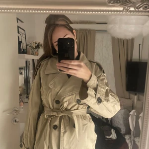 Beige trenchcoat med bälte - Säljer en beige trenchcoat med klassisk dubbelknäppning och bälte i midjan. Jackan har stora kragar, axelklaffar och långa ärmar med knappar vid mudden. Perfekt för dig som gillar stilrena och tidlösa plagg med snygg passform.