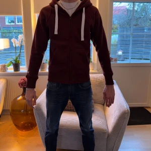 Vinröd hoodie med dragkedja - Nypris 799kr! Säljer en vinröd hoodie med vit snörning och dragkedja framtill. Tröjan har huva, långa ärmar och ribbade muddar. Perfekt för dig som gillar en enkel och stilren look. Passar till jeans eller joggers.