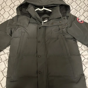 Svart dunjacka från Canada Goose - Säljer en svart dunjacka från Canada Goose med huva och klassisk logga på ärmen. Jackan har dragkedja och knappar framtill, samt två stora fickor på sidorna. Perfekt för kalla dagar och har en stilren look som funkar till det mesta.