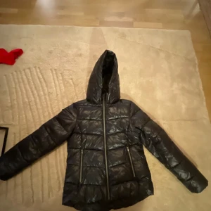 Svart pufferjacka från Everest - Svart pufferjacka från Everest med huva och två dragkedjeförsedda fickor. Jackan har quiltad design och är tillverkad i glansig polyester. Perfekt för kalla dagar och har en snygg, lite loose passform. Dragkedja framtill och stilren look. Den har även en liten repa 