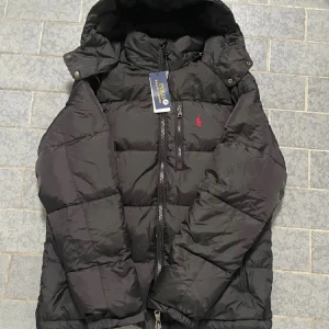 Svart pufferjacka Polo Ralph Lauren - Svart pufferjacka från Polo Ralph Lauren med huva och flera dragkedjefickor. Klassisk röd logga på bröstet och snygga detaljer som grova dragkedjor och quiltad design. Perfekt för kalla dagar och har en modern, loose passform.