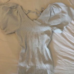 Vit ribbad topp med puffärm - Säljer en vit ribbad topp med puffiga korta ärmar och v-ringning. Toppen har knytband i ryggen och är figurnära i modellen. Materialet är stickat och ger en mjuk känsla. Perfekt för dig som gillar trendiga detaljer och vill ha en unik look.