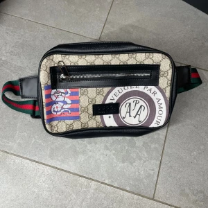  axelväska med tryck och dragkedja - Snygg axelväska med beige och svart mönster, coola tryck och detaljer i lila, blått och rött. Väskan har en framficka med dragkedja och bredt, randigt band i grönt och rött. Materialet är skinn och syntet, perfekt för att lyfta din outfit.