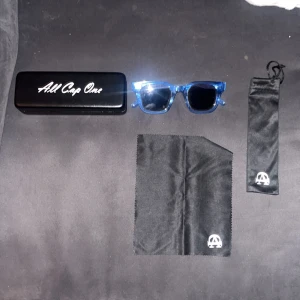 Blå transparenta solglasögon All Cap One - Snygga solglasögon från All Cap One med transparent blå båge och svarta glas. Rektangulär form som ger en cool vibe. Kommer med hårt fodral, mjuk påse och putsduk med logga. Perfekta för att sticka ut i mängden.