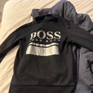 Svart tröja med silvertryck från Hugo Boss - Snygg svart tröja från Hugo Boss med stort silverfärgat BOSS-tryck och en bred silverrand på bröstet. Tröjan har lång ärm och ribbad nederkant. Perfekt för dig som gillar stilrena och moderna plagg med tydlig logga.