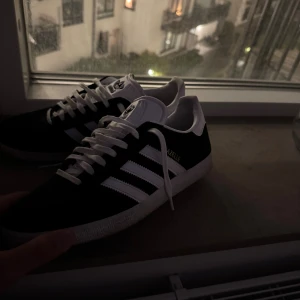 Adidas Gazelle svartvita sneakers - Säljer ett par klassiska Adidas Gazelle sneakers i svart mocka med vita detaljer och de ikoniska tre  ränderna på sidorna. Skorna har vit platt sula, snörning och Adidas-logga på plösen. Perfekta för dig som gillar en clean och tidlös stil som inte behövs nått utmärkt att matcha till!