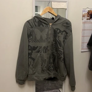Grå hoodie med grafiskt tryck - Säljer en grå hoodie med coolt svart grafiskt tryck framtill. Tröjan har dragkedja, huva och känguruficka. Mjuk insida och långärmad modell, perfekt för chill dagar. Passar dig som gillar streetwear vibes. Finns en vit fläck på vänstra ärmen som jag tror ska gå och få bort.